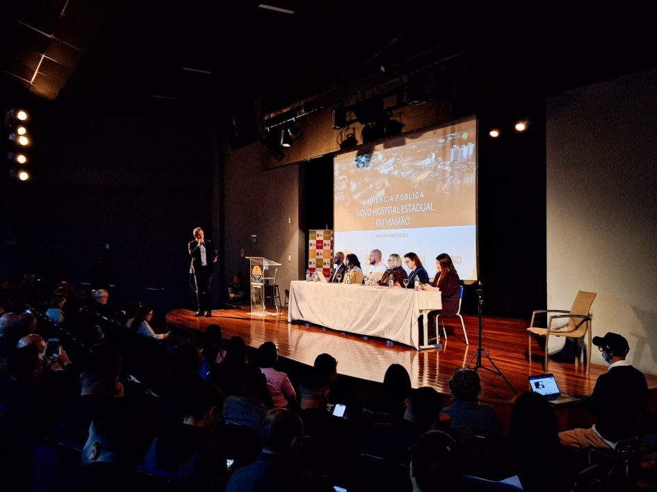 A imagem mostra uma audiência pública realizada em um auditório. No palco, há uma mesa comprida coberta com uma toalha branca, onde estão sentadas sete pessoas, aparentemente autoridades ou especialistas, participando de um debate ou apresentação. À esquerda do palco, um homem de terno está em pé, falando ao microfone próximo a um púlpito com o logotipo da prefeitura ou de algum órgão público.
Ao fundo, há um telão projetando uma apresentação com o título:
"Audiência Pública - Novo Hospital Estadual em Viamão"
O auditório está cheio, com várias pessoas sentadas assistindo à apresentação. À direita do palco, há uma cadeira vazia e uma pessoa sentada no chão com um notebook, aparentemente registrando ou transmitindo o evento. A iluminação do palco está concentrada nos participantes da mesa e no apresentador, enquanto o público está em ambiente mais escuro.